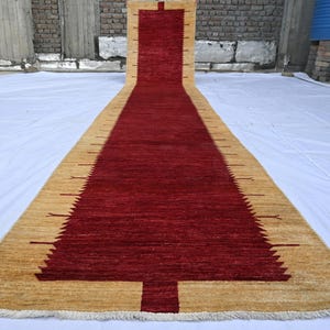 May include: A long, rectangular rug with a central red design and a tan border. The rug's central design resembles a stylized tree. The rug is made of a woven material and is placed on a white surface.