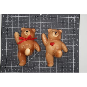 May include: Two light brown ceramic teddy bear wall plaques positioned on a black cutting mat with white grid lines. The left bear features a red ribbon tied around its neck, while the right bear has a small red heart detail on its chest.