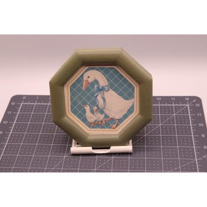 May include: An octagonal sage green frame surrounds a central illustration of a white goose wearing a blue ribbon, accompanied by two small goslings. The image is set against a blue grid background, positioned on a cutting mat with inch markings.