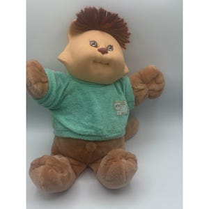 Peut inclure: Une peluche vintage avec un visage d'ours, des cheveux bruns et une chemise vert clair. Le jouet a des pattes marron et un corps marron. Un petit dessin de chat est sur la chemise.