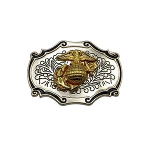 Puede incluir: Hebilla de cinturón plateada con un emblema dorado que representa un águila, un globo terráqueo y un ancla. La hebilla tiene un borde negro y un patrón ornamental en espiral. El emblema está centrado en la hebilla.
