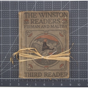 May include: An antique book titled "The Winston Readers Third Reader" with a decorative cover featuring a bird illustration. The book is bound with a raffia tie. The cover is a muted gray-green color.