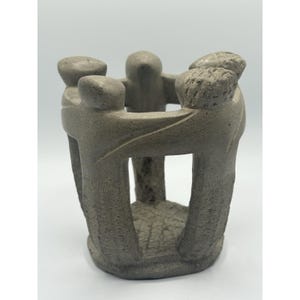 May include: A gray stone sculpture depicting four figures standing in a circle with their arms linked around each other's shoulders. The figures are carved from a single piece of textured stone, forming a hollow cylindrical structure.