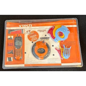 May include: A VTech V-Mix cordless phone set in original plastic packaging. The set includes an orange and black handset, a circular orange base, and several interchangeable colourful plastic faceplates and base covers in red, yellow, and blue.
