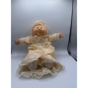 May include: A vintage Cabbage Patch Kid doll wearing a cream-colored dress with lace trim and a matching bonnet. The doll has a pacifier in its mouth and is in a seated position with its arms outstretched. The doll's skin is a peach color.