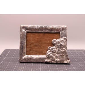May include: A rectangular silver-toned metal picture frame with a textured, bark-like border. The frame features a raised, sculpted design of two teddy bears in the bottom right corner. The centre of the frame is a plain, dark brown wood insert.