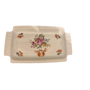 Könnte beinhalten: Rechteckige, cremefarbene Keramikservierplatte mit floralem Design. Die Platte hat einen zentralen Strauß aus rosa, lila und gelben Blumen mit kleineren floralen Akzenten. Goldfarbene Zierleisten säumen die Platte und die Griffe.