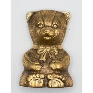 May include: A brass-coloured teddy bear figurine with a bow tie. The bear is sitting with its paws visible and has detailed fur texture. The figurine is on a white background.