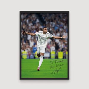 May include: Framed print featuring a soccer player in a white uniform with black accents, celebrating on a green field. The image includes the text "To Name Best Wishes" and a signature. The background shows a crowd of spectators.