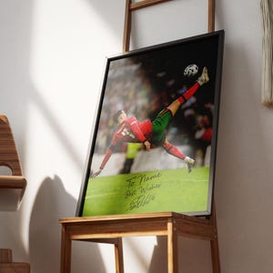 Puede incluir: Impresi&oacute;n enmarcada de un futbolista en el aire, pateando el bal&oacute;n. El jugador viste un uniforme rojo y verde. La impresi&oacute;n incluye el texto "To Name Best Wishes" y una firma. El marco es negro.
