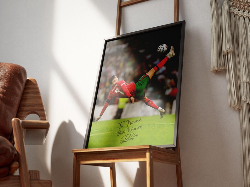 Puede incluir: Impresi&oacute;n enmarcada de un futbolista en el aire, pateando el bal&oacute;n. El jugador viste un uniforme rojo y verde. La impresi&oacute;n incluye el texto "To Name Best Wishes" y una firma. El marco es negro.