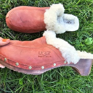 May include: Reddish-brown UGG Australia clogs with wooden heels and sheepskin lining. The clogs have a studded trim. The image shows the clogs on a bed of green grass.