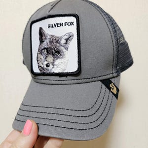 May include: A grey trucker cap featuring a square patch on the front with a detailed embroidered illustration of a fox and the text SILVER FOX. The cap has a solid grey front panel, a mesh back, and a small black tab with a gold icon on the brim.