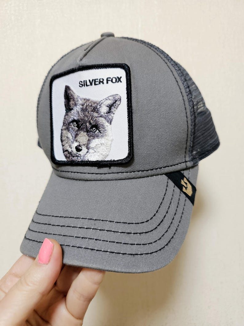 May include: A grey trucker cap featuring a square patch on the front with a detailed embroidered illustration of a fox and the text SILVER FOX. The cap has a solid grey front panel, a mesh back, and a small black tab with a gold icon on the brim.