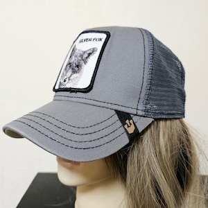May include: A grey trucker hat featuring a mesh back and a front patch with a graphic illustration of a fox and the text SILVER FOX. The brim has black stitching and a small black tab with a gold-colored icon on the side of the visor.