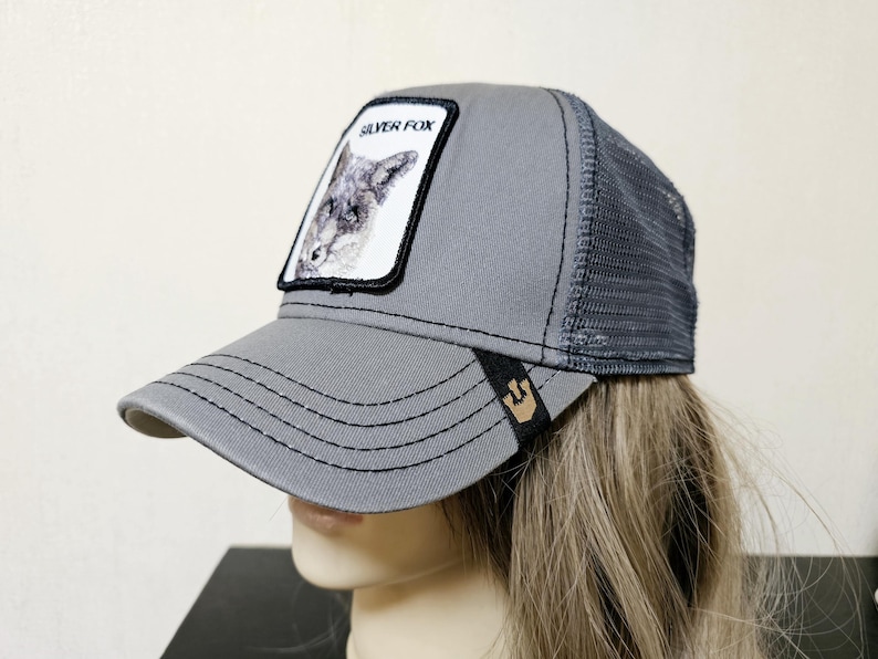 May include: A grey trucker hat featuring a mesh back and a front patch with a graphic illustration of a fox and the text SILVER FOX. The brim has black stitching and a small black tab with a gold-colored icon on the side of the visor.