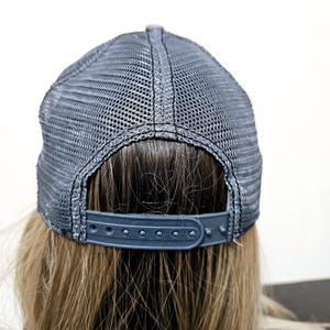 May include: A dark gray trucker hat featuring a mesh back panel and an adjustable plastic snapback closure. The cap is constructed with a structured crown and a breathable mesh design, providing a secure fit with its multi-hole strap mechanism.
