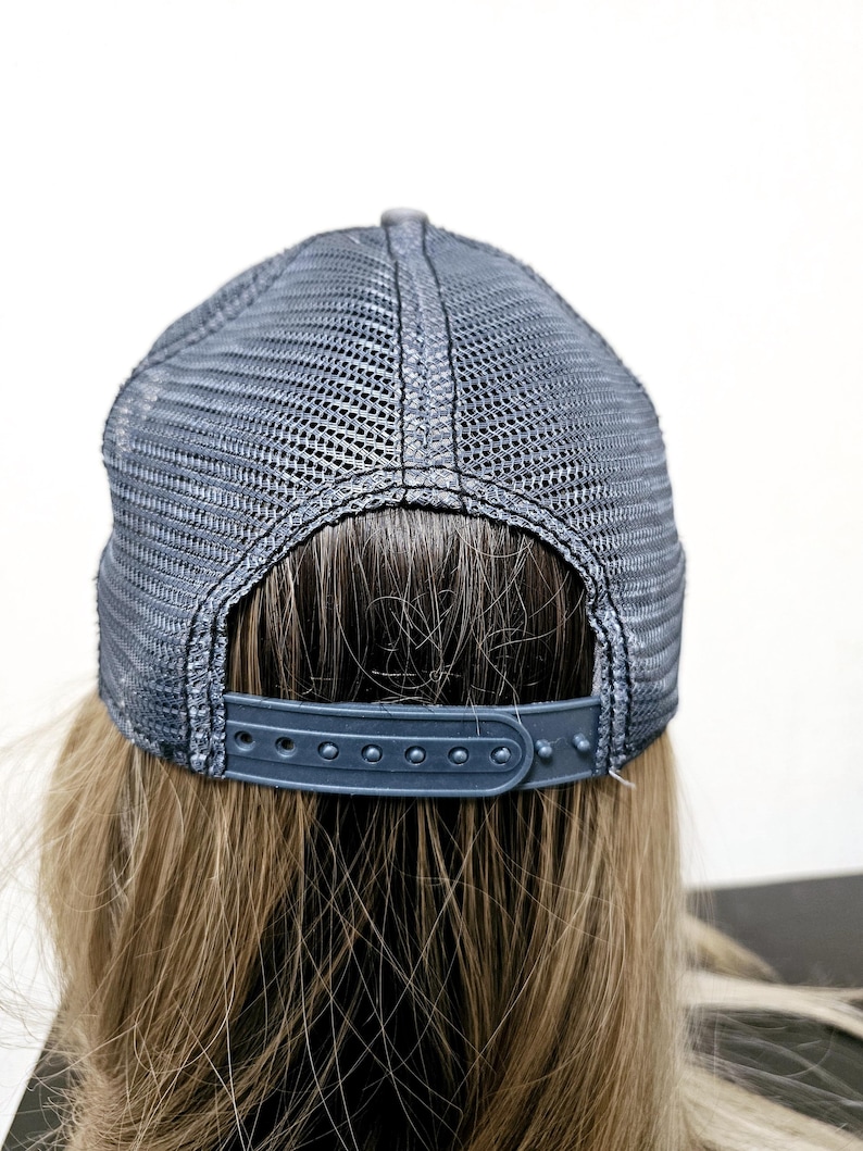 May include: A dark gray trucker hat featuring a mesh back panel and an adjustable plastic snapback closure. The cap is constructed with a structured crown and a breathable mesh design, providing a secure fit with its multi-hole strap mechanism.