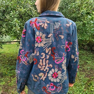 May include: A denim jacket with intricate floral embroidery in shades of pink, red, and beige. The jacket is a dark blue color and has a button-down front. The embroidery is detailed and covers the back and sleeves of the jacket.