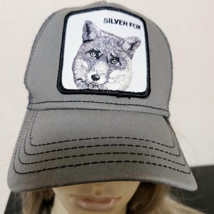 May include: A grey trucker hat featuring a white rectangular patch on the front panel. The patch displays an embroidered illustration of a fox head with the text SILVER FOX printed in black above the animal. The hat has a curved brim with stitching.