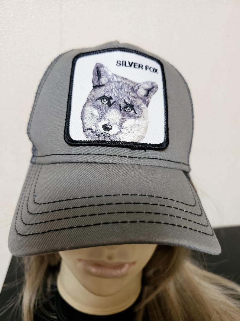 May include: A grey trucker hat featuring a white rectangular patch on the front panel. The patch displays an embroidered illustration of a fox head with the text SILVER FOX printed in black above the animal. The hat has a curved brim with stitching.