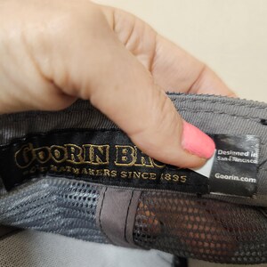 May include: A grey baseball cap interior featuring a black fabric label with gold text reading Goorin Bros. Hatmakers Since 1895. A smaller white and black tag attached to the side displays the text Designed in San Francisco and Goorin.com.