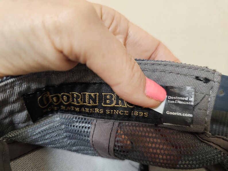 May include: A grey baseball cap interior featuring a black fabric label with gold text reading Goorin Bros. Hatmakers Since 1895. A smaller white and black tag attached to the side displays the text Designed in San Francisco and Goorin.com.