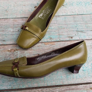 May include: A pair of olive green leather pumps with a square toe and a small heel. The shoes feature a decorative strap with a buckle. They are displayed on a weathered, light blue and brown wooden surface. The brand name is visible inside one shoe.