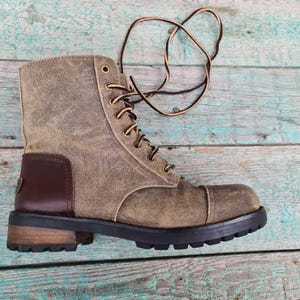 Könnte beinhalten: Ein verwitterter brauner Lederstiefel mit Schnürdesign. Der Stiefel hat einen dunkelbraunen Lederaufnäher an der Seite und eine schwarze Sohle. Die Schnürsenkel sind braun und schwarz. Der Stiefel steht vor einem verwitterten, türkisfarbenen Holzhintergrund.