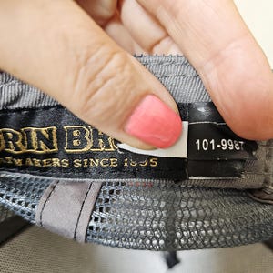 May include: A grey baseball cap interior featuring a black fabric label with gold embroidered text reading "GOORIN BROS" and "HATMAKERS SINCE 1895". A smaller black tag attached to the right displays the white printed numerical code 101-9987.