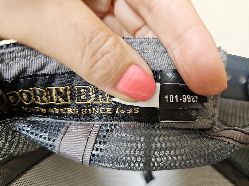 May include: A grey baseball cap interior featuring a black fabric label with gold embroidered text reading "GOORIN BROS" and "HATMAKERS SINCE 1895". A smaller black tag attached to the right displays the white printed numerical code 101-9987.