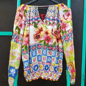 May include: A long-sleeved blouse with a vibrant floral pattern in pink, red, yellow, and blue. The blouse features a keyhole back and is hanging on a black hanger against a dark background. The sleeves and body are covered in a repeating floral design.