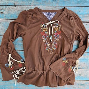 May include: Brown long-sleeved blouse with floral and butterfly embroidery. The blouse has a V-neck with a tie, a ruffled hem, and bell sleeves with embroidered details. The fabric is soft and the blouse is a bohemian style.