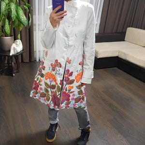May include: A white double-breasted coat with a high collar and floral design. The coat features a colorful floral pattern with orange, pink, and purple flowers. The coat is worn with grey jeans and black boots.