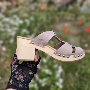 Könnte beinhalten: Eine hellgraue Clog-Sandale mit einem Blockabsatz aus hellem Holz. Der obere Riemen ist mit dem UGG-Logo geprägt und mit kleinen, runden, goldfarbenen Metallnieten an den Seiten an der Holzsohle befestigt.