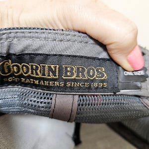 May include: A grey baseball cap featuring an interior black fabric label with gold embroidered text that reads Goorin Bros. Bold Hatmakers Since 1895. A smaller black tag attached to the side of the label displays the text One Size in white.