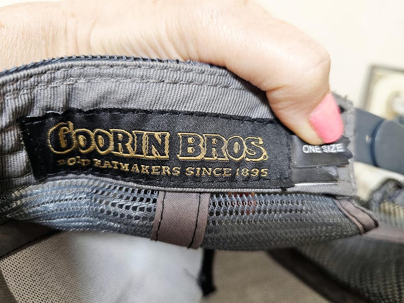 May include: A grey baseball cap featuring an interior black fabric label with gold embroidered text that reads Goorin Bros. Bold Hatmakers Since 1895. A smaller black tag attached to the side of the label displays the text One Size in white.