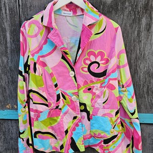 Puede incluir: Una vibrante blazer rosa con un colorido estampado abstracto en tonos verdes, azules y negros. La blazer tiene un cuello clásico, dos bolsillos delanteros y dos botones blancos. La etiqueta dice "Pierre Cardin".