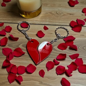 May include: Two red heart-shaped keyrings, each half engraved with "Him" and "Her", connected by a chain and keyring. The keyrings are surrounded by red rose petals on a wooden surface. A candle in a glass holder is in the background.