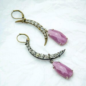 May include: A pair of crescent-shaped earrings with clear gemstones and a dangling pink gemstone. The earrings have a vintage aesthetic with a gold-tone metal and are set against a white background. The pink gemstones are a light, dusty rose color.