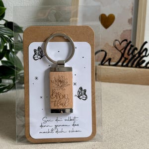 Könnte beinhalten: Ein Holzschlüsselanhänger mit einem silberfarbenen Metallring. Der Holzteil ist mit einer Lotusblume und den Worten "Be You tiful" graviert. Der Schlüsselanhänger ist mit einer Karte mit Schmetterlingsillustrationen und Text auf Deutsch verpackt.