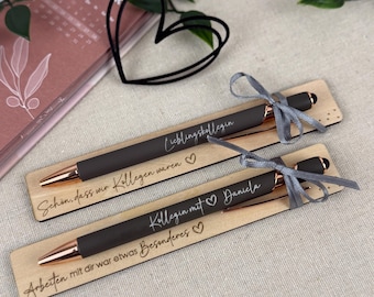 May include: Two black pens with rose gold accents and gray ribbons, presented on wooden holders. The pens have text that reads "Lieblingskollegin" and "Kollegin mit Daniela". A calendar and a wire heart are in the background.