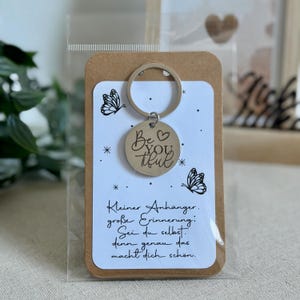 Könnte beinhalten: Ein silberfarbener Schlüsselanhänger mit einem runden Anhänger mit der Aufschrift "Be YOU tiful". Der Schlüsselanhänger ist an einer Karte mit Schmetterlingsillustrationen und deutschem Text befestigt. Die Karte ist in einer transparenten Plastikhülle verpackt.