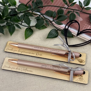 Könnte beinhalten: Zwei roségoldene Stifte mit goldenen Akzenten und schwarzen Stiftspitzen, präsentiert auf Holzplatten. Der obere Stift trägt die Aufschrift "Cheun mit Daniela" und "Danke für die gemeinsame Reise."