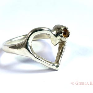 May include: A silver ring in the shape of a heart, with a small, round, brown gemstone set at the top. The ring is on a white background, with the artist's name, Gisela Backe, in the bottom right corner.
