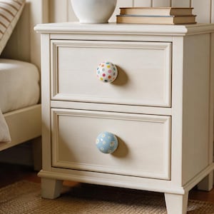 May include: A cream-coloured wooden bedside table with two drawers, featuring a top drawer knob with a multicoloured polka dot pattern and a bottom drawer knob with a light blue floral pattern. The bedside table has a rectangular frame and four legs.