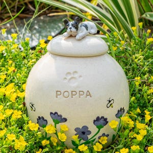May include: A cream ceramic pet urn with a lid, topped with a small dog figurine. The urn is decorated with paw prints, floral designs, and the name "POPA". The urn is surrounded by yellow flowers and greenery, creating a garden setting.