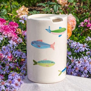 May include: A cylindrical, off-white ceramic container with a lid and a cutout. The container is decorated with colourful illustrations of fish in shades of blue, green, and pink. The background features a variety of flowers.