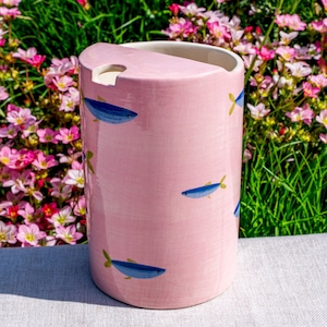 May include: A cylindrical ceramic utensil holder with a glossy light pink finish, decorated with small blue fish illustrations. The top edge features a semi-circular cutout and a small notch, revealing a white interior and a smooth, glazed surface.
