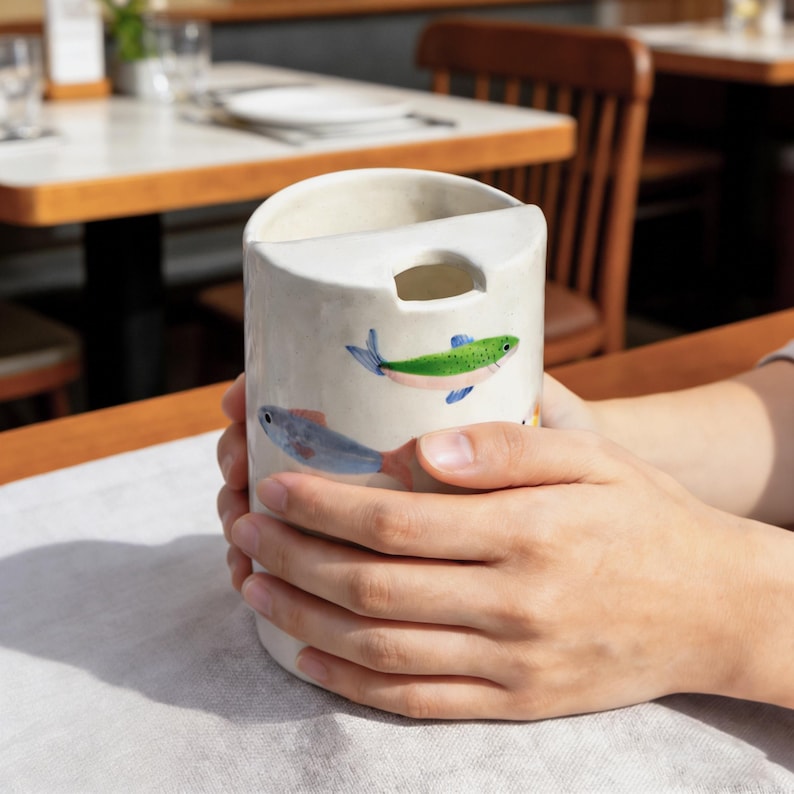 May include: A cylindrical ceramic cup with a white base and a unique top opening featuring a cutout notch. The exterior is decorated with hand-painted illustrations of small fish in shades of green, blue, and orange, arranged around the body.